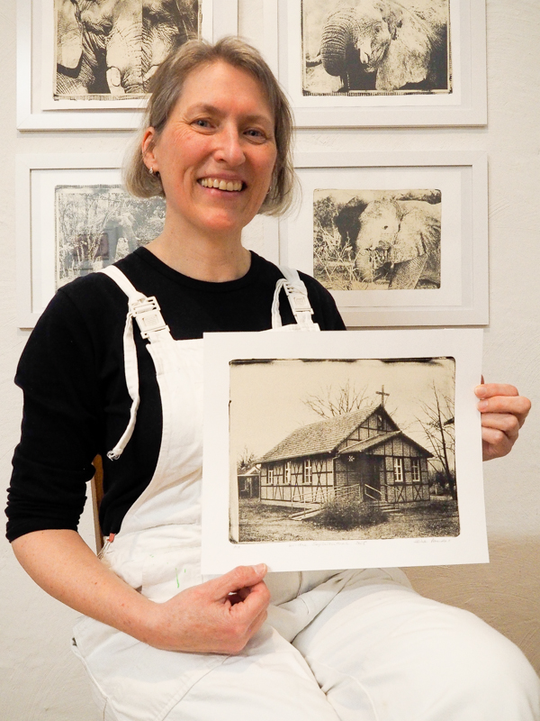 Heike Pander im Atelier, 2025 Das Bild zeigt ein Porträt der Künstlerin Heike Pander in ihrem Atelier. Sie sitzt auf einem Stuhl und hält einen Gel-Druck in die Kamera. Auf ihm ist das Abbild einer kleinen Kirche im Oderbruch zu sehen. Im Hintergrund sind Bilder von Elefanten zu sehen. Alle Bilder sind gedruckte Fotounikate von der Gel-Platte.