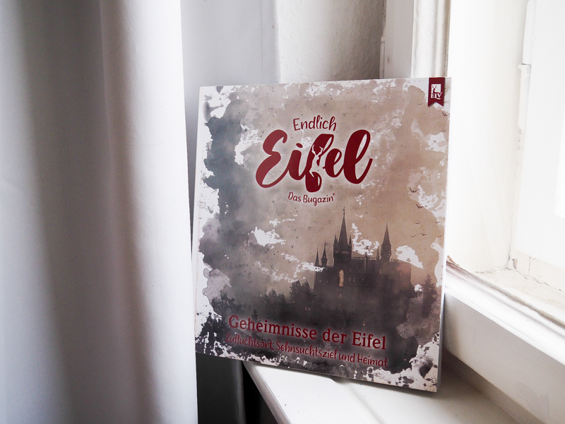 Das Bild zeigt ein Foto des Buchs "Endlich Eifel. Das Bugazin", an dem Heike Pander als Co-Autorin mitgewirkt hat. Auf dem Umschlag sind Titel des Buchs, Untertitel und Verlag in roter Schrift zu sehen, sowie eine düster wirkende Burg, die aquarelliert wirkt. Das Buch steht vor einem Fenster und lehnt gegen den Fensterrahmen.