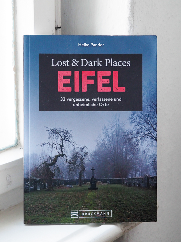 Das Bild zeigt ein Foto des Buchs "Lost & Dark Places Eifel" der Autorin Heike Pander . Das Buch steht vor einem Fenster und lehnt gegen den Fensterrahmen. Auf dem Cover ist der Titel in weißer und roter Schrift abgebildet, der Name des Verlags in weißer Schrift sowie ein Foto des Tuchmacherfriedhofs in Menzerath bei Monschau.