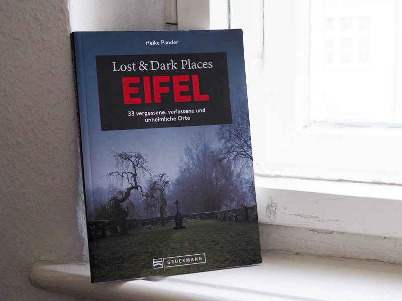 Das Bild zeigt ein Foto des Buchs "Lost & Dark Places Eifel" der Autorin Heike Pander . Das Buch steht vor einem Fenster und lehnt gegen den Fensterrahmen. Auf dem Cover ist der Titel in weißer und roter Schrift abgebildet, der Name des Verlags in weißer Schrift sowie ein Foto des Tuchmacherfriedhofs in Menzerath bei Monschau.