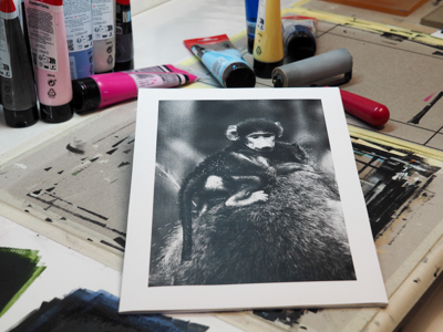 Fotovorlage Das Foto zeigt einen Arbeitsplatz zur Herstellung einer Monotypie. Zu sehen sind verschiedene Acryl-Farben, eine Walze zum Ausrollen der Farben, sowie ein schwarz-weiß ausgedrucktes Foto im Format A4, das im Prozess als Druckstock dienen wird. Auf dem Foto ist ein kleiner Pavian zu sehen.