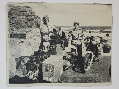 Das Bild zeigt eine Monotypie, einen Gel-Druck von einer Fotografie. Zu sehen sind Heike Pander und Matthias Kuhlmann in Motorradbekleidung, vor ihnen stehen ihre Reisemotorräder. Das Bild wurde am Kap der Guten Hoffnung nach einer mehrmonatigen Motorradreise durch das südliche Afrika aufgenommen.